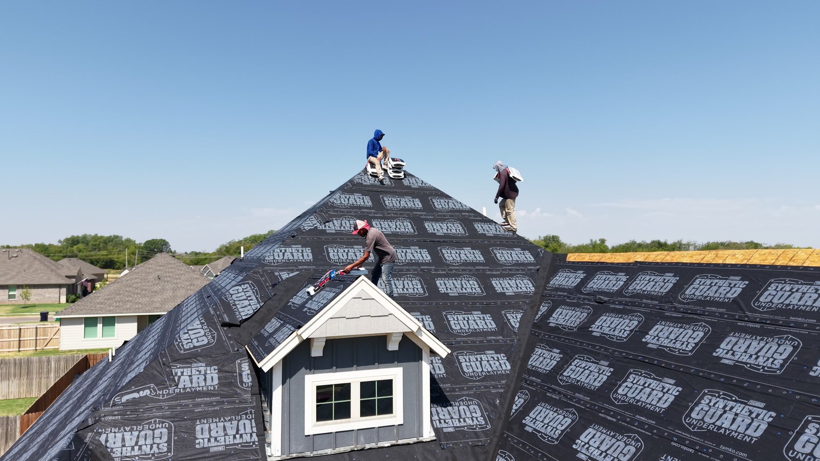 Three roofers working near the ridge of a hip roof covered in black synthetic underlayment under a clear sky